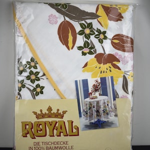 May include: A packaged white tablecloth with a floral pattern in brown, yellow, and green. The packaging features a label with the word "ROYAL" and the text "DIE TISCHDECKE IN 100% BAUMWOLLE". A smaller image shows the tablecloth on a table.