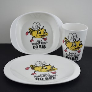 May include: A white plastic dinnerware set consisting of a bowl, a plate, and a cup. Each piece features a graphic of a yellow and black bee with the text I AM A ROMPER ROOM DO BEE printed in black letters below the illustration of the cartoon bee.