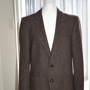 May include: A brown tweed blazer with two front pockets and two buttons. The blazer is displayed on a white mannequin. The background includes a window with white blinds and a glimpse of an outdoor scene.