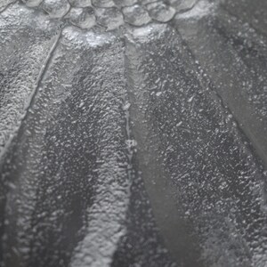 May include: A clear glass plate featuring a raised, textured sunflower petal design. The surface has a pebbled, frosted appearance with smooth, defined lines radiating from a central cluster of small, rounded glass beads at the top.