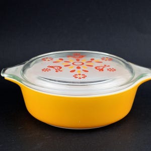May include: A vintage, round, yellow casserole dish with a clear glass lid. The lid features a floral and bird design in orange and yellow. The dish has two handles and is set against a black background.