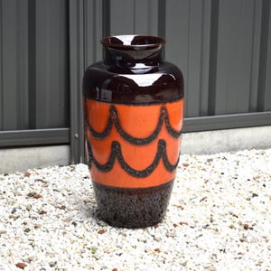 May include: A large ceramic vase with a glossy finish. The top and bottom sections are dark brown, while the middle section is orange with a repeating black wave pattern. The vase is standing on a bed of small, white pebbles.