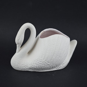 May include: A white ceramic swan-shaped planter with a textured finish. The swan features a curved neck and a wide opening for plants or decorative items. The planter is set against a dark background.