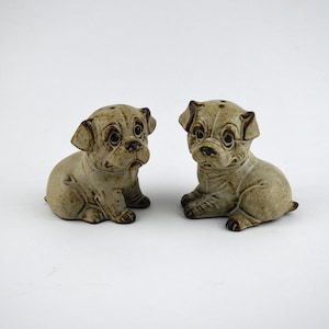 May include: Two ceramic bulldog figurines in a seated position. The dogs are light brown with darker brown accents, including their eyes, ears, and paws. The salt and pepper shakers have a matte finish and are set against a white background.