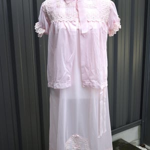 May include: A light pink nightgown set featuring a short-sleeved top with a Peter Pan collar, a ribbon tie at the neck, and lace trim on the sleeves and bodice. The matching skirt has a sheer overlay with a decorative lace panel at the hem.
