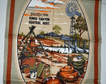 May include: A tan tea towel with a red border features a colorful illustration of an outback scene in Australia. The scene includes a campfire, a windmill, a bridge, and a sign that reads "Greetings from Kings Canyon Central Aust."