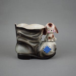 May include: A ceramic boot-shaped planter in shades of gray and black, with a small, brown and white puppy figurine perched on the side. The boot has a blue patch detail, and the puppy has closed eyes.