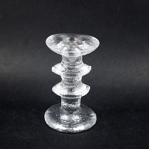 May include: A clear glass candle holder with a textured surface. The candle holder has a round base, a central column with three stacked tiers, and a flat top. The design is simple and elegant, suitable for various decor styles.
