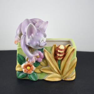 May include: A ceramic planter featuring a lavender-colored cat perched on the edge. The planter is rectangular with a green and yellow floral design, including flowers and leaves. The cat has a smiling expression.