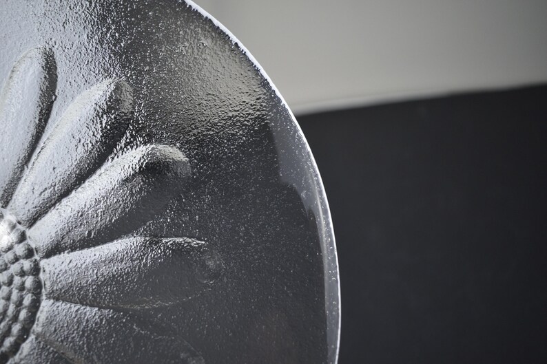 May include: A clear glass plate featuring a raised, textured floral design on its underside. The embossed pattern depicts a central flower with numerous radiating petals, creating a tactile surface that catches the light across the circular edge.
