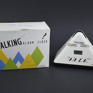 May include: A white and black Talking Alarm Clock with a pyramid shape, next to its box. The clock displays the time 2:01. The box has the words "TALKING ALARM CLOCK" and "T-10 junior" printed on it.