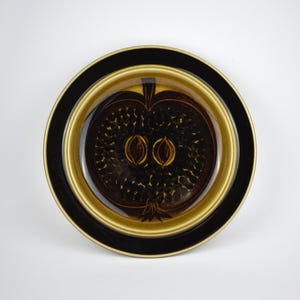 May include: A round ceramic plate with a black rim and a golden-brown inner ring. The center features a dark brown apple design with two seed shapes. The plate is likely used for serving food.
