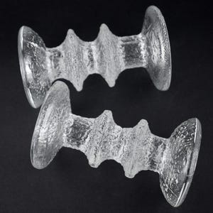 May include: Two clear glass candle holders with a textured, ridged design. Each holder has a disc-shaped base and top, connected by a central, textured column. The glass has a slightly frosted appearance, and the background is black.