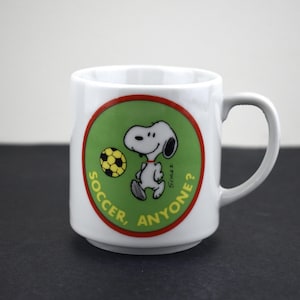 May include: White ceramic mug featuring a cartoon Snoopy character with a soccer ball. The mug has a green and red circular design with the words "SOCCER, ANYONE?" printed around the image. The mug has a gray handle.