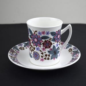 May include: White teacup and saucer set with a floral pattern in shades of purple, pink, and brown. The cup has a curved handle and sits on a matching saucer. The floral design is repeated on the saucer's rim.