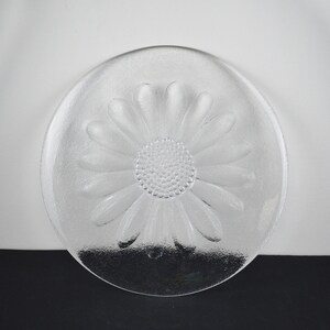 May include: A clear glass plate featuring a molded daisy flower design. The center of the plate displays a textured, dotted pattern representing the flower head, surrounded by smooth, elongated petals that extend toward the circular rim.