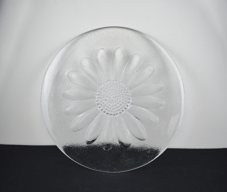May include: A clear glass plate featuring a molded daisy flower design. The center of the plate displays a textured, dotted pattern representing the flower head, surrounded by smooth, elongated petals that extend toward the circular rim.