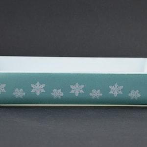 May include: A rectangular teal baking dish with white snowflake designs. The dish has a white interior and white trim around the rim. The dish is approximately 30 cm long.