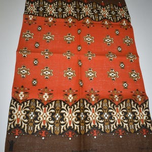 May include: An orange and brown patterned textile with a repeating design of stylized floral and geometric shapes. The textile has a dark brown border at the top and bottom, with the words "Save the Children" visible at the bottom.