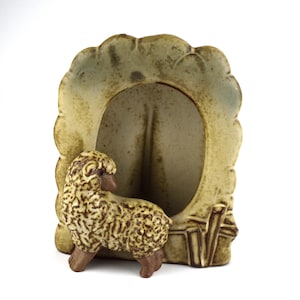 以下が含まれることがあります： 羊の置物付きのセラミックフォトフレーム。フレームは薄緑色で、スカラップエッジが付いています。羊は茶色とタン色で、ウールのような質感があります。フレームには垂直の開口部があります。