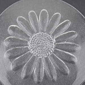 May include: A circular clear glass plate featuring a raised, embossed daisy flower design in the center. The flower has a textured, dotted circular core surrounded by numerous smooth, elongated petals that extend outward toward the plate edge.