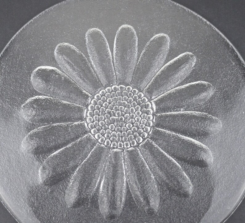 May include: A circular clear glass plate featuring a raised, embossed daisy flower design in the center. The flower has a textured, dotted circular core surrounded by numerous smooth, elongated petals that extend outward toward the plate edge.