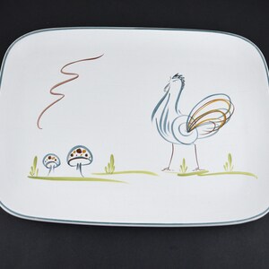 May include: A rectangular white ceramic serving platter with rounded corners and a thin blue rim. The surface features a stylised illustration of a rooster in blue and brown, two small mushrooms with blue caps, and green grass blades.
