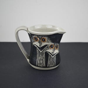 May include: A ceramic jug with a handle, featuring a black body adorned with a white owl face design. The owl's eyes are brown. The rim and handle are light grey. The jug is likely used for serving beverages.