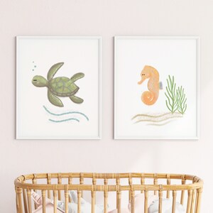 Two framed illustrations of a sea turtle and a sea horse on the wall above a crib in a baby girl’s nursery