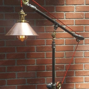 Op de afbeelding: Industriële vloerlamp met een koperen kap en zichtbare gloeilamp. De lamp heeft zwarte buizen, messing fittingen en een rood snoer. Het ontwerp bevat een katrolsysteem en staat tegen een bakstenen muur. De tekst "Brightsideoflights" is zichtbaar.