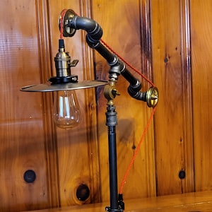 以下が含まれることがあります： 赤いコードと真鍮の滑車が付いた、黒い金属製の steampunk スタイルのデスクランプ。ランプは黒い金属製のシェードと透明なガラスの電球が付いています。