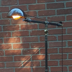 Peut inclure: Lampadaire de style industriel avec une structure en tuyaux noirs et un abat-jour en dôme en métal foncé. La lampe est équipée d'une ampoule chaude et brillante et d'un cordon décoratif. La lampe est placée devant un mur de briques rouges.