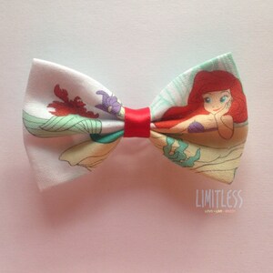 May include: A red, white, and green bow tie with a printed image of Ariel, the Little Mermaid, from the Disney movie. The bow tie has a red ribbon in the center.