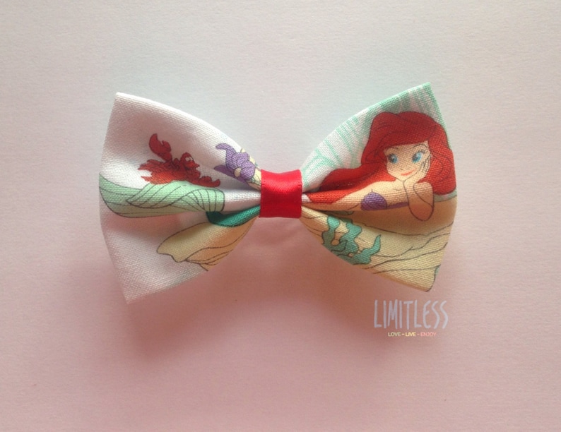 May include: A red, white, and green bow tie with a printed image of Ariel, the Little Mermaid, from the Disney movie. The bow tie has a red ribbon in the center.
