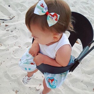 May include: A baby girl wearing a white tank top and a blue and white skirt with a mermaid print. She is wearing a red and white bow with a mermaid print in her hair. She is sitting in a black and gray baby swing.