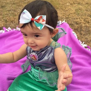 May include: A baby girl wearing a green and purple mermaid costume with a headband featuring a white bow with a red center and a printed image of Ariel from The Little Mermaid.