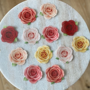 Puede incluir: Una variedad de recortes de rosas de papel en varios colores, incluyendo rojo, rosa y amarillo, con hojas verdes. Las rosas están dispuestas sobre una superficie de mármol blanco, mostrando su diseño detallado y apariencia realista.