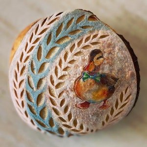 Może przedstawiać: Okrągły bochenek chleba z niebiesko-białym wzorem liści i namalowanym obrazem kaczki w muszce.