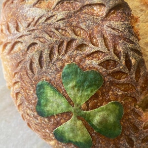 May include: A close-up of a round loaf of bread with a green shamrock design. The bread is brown and has a rough texture.