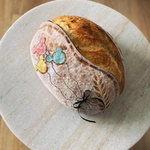 May include: A loaf of sourdough bread with a hand-drawn design of Mickey Mouse characters holding balloons. The bread is on a white marble surface.