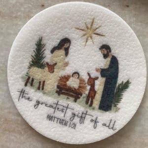 May include: A white round ornament with a nativity scene illustration. The illustration features Mary, Joseph, and baby Jesus in a manger with a star above them. The text "the greatest gift of all" and "Matthew 1:21" is printed below the illustration.