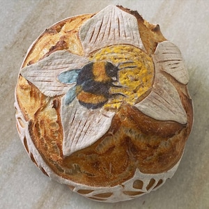May include: A round loaf of sourdough bread with a hand-painted design of a bee and flower. The bee is yellow and black, and the flower is white with yellow accents.