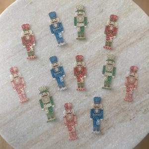 May include: A selection of miniature nutcracker figurines in shades of red, blue, green, and pink. Each figurine features a detailed design with a hat, uniform, and mustache. The figurines are displayed on a light-coloured, speckled surface.