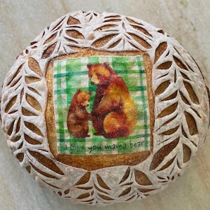 May include: A white sourdough bread loaf with a decorative design of leaves. The loaf is decorated with a printed image of two bears and the text "I love you mama bear".