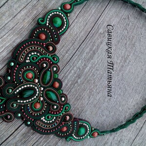 May include: A green, brown and red beaded necklace with a large, intricate design. The necklace is made of multiple strands of beads and features a variety of shapes and sizes. The necklace is on a wooden surface.
