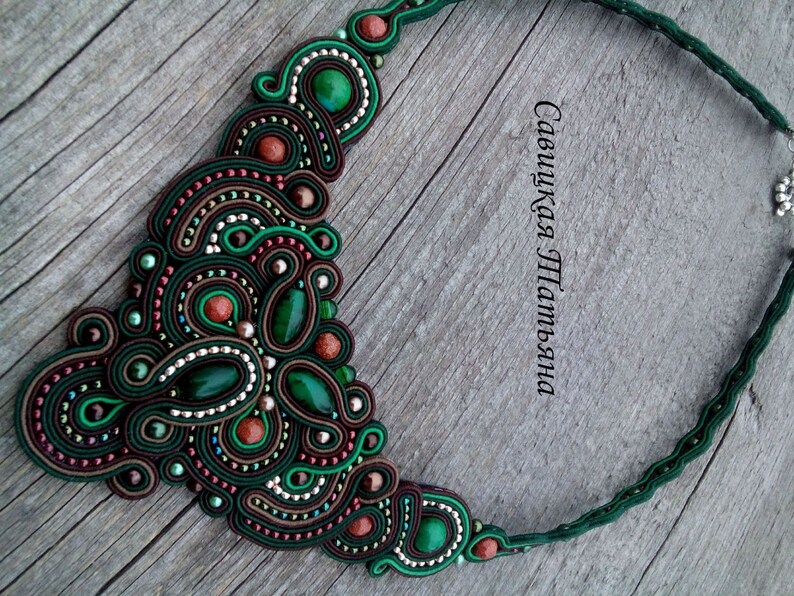 May include: A green, brown and red beaded necklace with a large, intricate design. The necklace is made of multiple strands of beads and features a variety of shapes and sizes. The necklace is on a wooden surface.