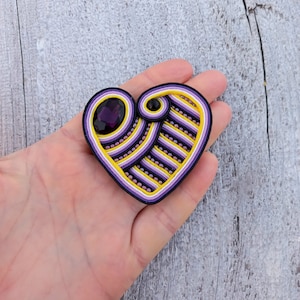 Op de afbeelding: Een hartvormige broche gemaakt van paarse, gele en witte draad met een paarse edelsteen in het midden.