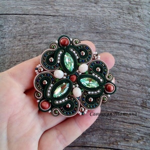 May include: A green, white, and brown beaded brooch with a large green crystal in the center. The brooch is held in a hand and the text "Савицкая Платвина" is visible in the bottom right corner.