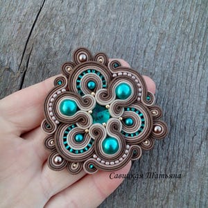 Op de afbeelding: Een bruine en beige kralen broche met een turquoise centrum en een groen en gouden ontwerp. De broche ligt op een houten oppervlak.
