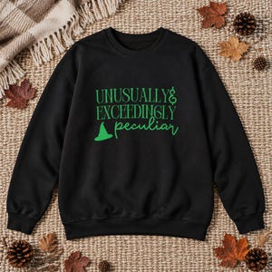 May include: A black long-sleeve sweatshirt featuring the green text "UNUSUALLY EXCEEDINGLY peculiar" printed on the front. The design includes a small green witch hat icon positioned below the word "EXCEEDINGLY" and to the left of "peculiar".